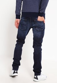 Jean en denim bleu foncé avec une coupe ajustée, cinq poches et un léger délavage. Associé à des baskets blanches et un pull en laine bleu marine.