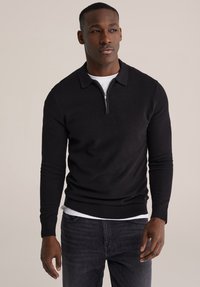 Črn pleten pulover z zadrgo na ovratniku, dolgimi rokavi, rebrastimi manšetami in spodnjim robom, nosi se čez belo srajco, združen s temnimi kavbojkami.