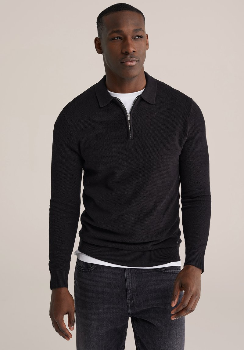 Črn pleten pulover z zadrgo na ovratniku, dolgimi rokavi, rebrastimi manšetami in spodnjim robom, nosi se čez belo srajco, združen s temnimi kavbojkami.