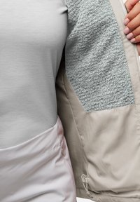 Lichtgrijze jas met zachte fleece voering in een contrasterend lichtgrijs gedeelte; ritsen en trekkoorden zichtbaar aan de onderkant.