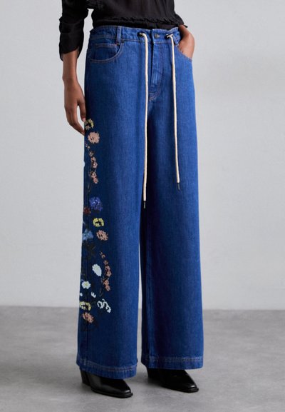 Wijdvallende denim broek in donkerblauw, met een bloemenborduursel aan de linkerkant en een trekkoord in de taille. Zwarte hakken.
