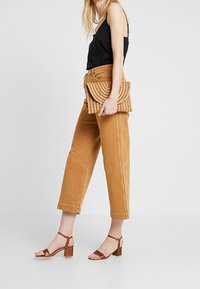 Kvinna i beige beskurna byxor, svart ärmlös topp, bruna klacksandaler, som håller en vävd beige clutchtväska mot en vit bakgrund.