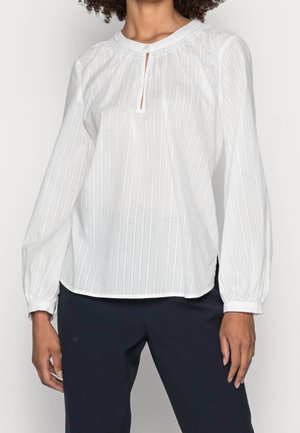 Femme portant un chemisier blanc à manches longues avec une texture à rayures verticales et un décolleté en trou de serrure, associée à un pantalon foncé.