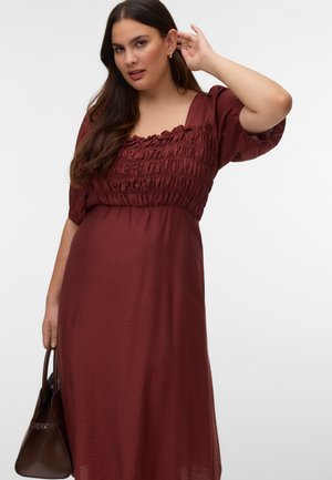 Femme en robe bordeaux texturée avec des manches bouffantes tient un sac à main marron dans une main et touche ses cheveux de l'autre.