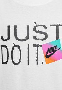 Camiseta blanca con texto en negrita negro que dice "SOLO HAZLO", con una pegatina del logotipo de Nike colorida en la esquina, mostrando un degradado de rosa y azul.