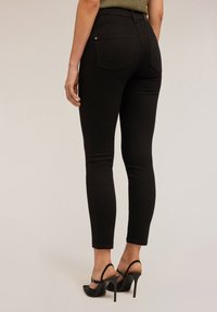 Mujer vestida con jeans ajustados negros y zapatos de tacón slinkback negros, de pie contra un fondo neutro liso.