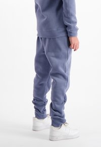 Persoon in blauwe joggerpants en een bijpassende sweatshirt met witte sneakers die tegen een witte achtergrond staat.