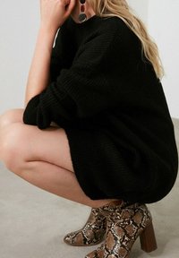 Femme accroupie portant une robe-pull noire en maille, de grandes boucles d'oreilles rondes et des bottines marron à talons avec un motif peau de serpent.