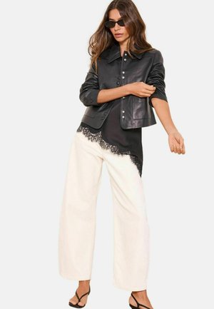 Woman wearing black leather jacket, black lace-trimmed top, white wide-leg pants, black sandals, and sunglasses, adjusting sleeve on light background.