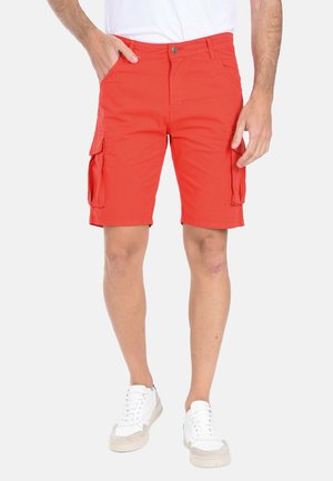 Uomo che indossa pantaloncini cargo arancioni vivaci con tasche, scarpe da ginnastica bianche e una maglietta bianca, in piedi davanti a uno sfondo semplice.