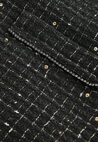 Juodas tweed audinys su auksinėmis blizgučiais ir balto siūlo detalėmis, pasižymintis tekstūruota paviršiumi ir lankstytu krašto dizainu.