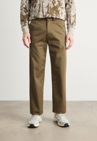 Brown cotton trousers with a straight-leg design, featuring a button closure and side pockets, paired with grey sneakers.