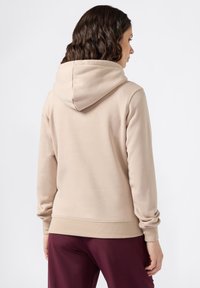 Hoodie beige con cappuccio regolabile, polsini e orlo a coste. Il tessuto appare morbido con una superficie liscia, indossato con pantaloni borgogna.