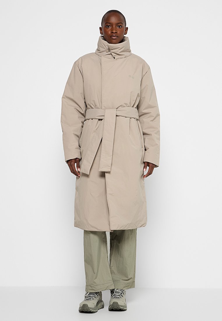 Beige pufferjack met hoge kraag, ceintuur in de taille, lange mouwen en zakken aan de zijkant; gedragen over lichtgroene broek en sneakers.