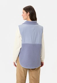 Hellblaues Weste aus einer Mischung aus glattem Stoff und flauschigem Fleece, mit einem Kragen und abgerundetem Saum, getragen über einem weißen Langarmhemd.