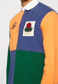 Mångfärgad rugbytröja med orange, lila och gröna paneler. Framsidan pryds av en broderad röd ros på en vit lapptäck.