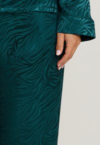 Tissu bleu-vert avec un motif de rayures zebra subtil ; texture lisse. Poignet de la manche visible et main reposant contre le tissu.