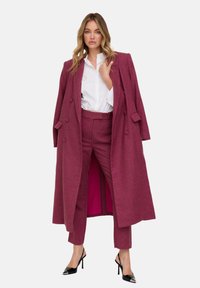 Mantel aus einer Wollmischung in Kastanienbraun mit doppelreihigem Design, kombiniert mit passenden, maßgeschneiderten Hosen und einem weißen Hemd mit Knöpfen. Schwarze High Heels vervollständigen den Look.