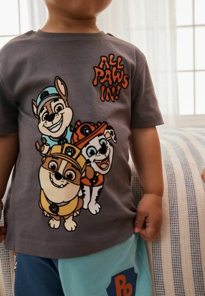 Kind trägt ein graues Hemd mit drei animierten Welpen in Bauhelmen und dem Text "Alle Pfoten rein!", steht drinnen neben einem gestreiften Kissen.