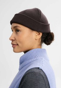 Bruine geribbelde gebreide beanie op het hoofd van een persoon, met een lichtblauw gebreid vest met hoge kraag dat zichtbaar is rond de hals.