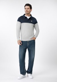 Γκρι και μπλε ναυτικό μακρυμάνικο πόλο με γιακά, με λογότυπο στο στήθος, συνδυασμένο με φαρδιά μπλε τζιν και λευκά αθλητικά παπούτσια.