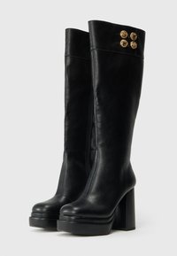 Črne čizme do kolen s platformo in robustnimi petami, z zlatimi okrasnimi gumbki blizu vrha in stranskim zadrgo.
