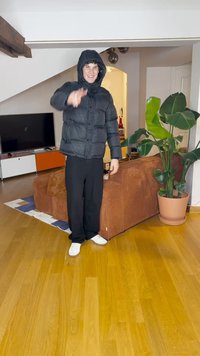 Giacca a vento nera con cappuccio, pantaloni neri larghi e scarpe bianche. In piedi su un pavimento in legno accanto a un divano marrone e a una pianta verde.