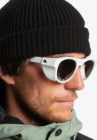 White sunglasses with thick frames and dark lenses. Worn with a black ribbed knit beanie and a green jacket, highlighting layered textures.