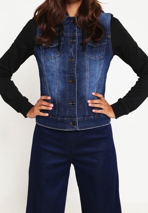 Personne portant un gilet en jean bleu avec des manches longues noires et un pantalon large bleu marine, debout les mains sur les hanches.