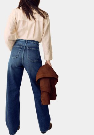 Femme en jean bleu taille haute à jambes larges et blouse crème tenant une veste marron pliée, debout devant un fond uni.