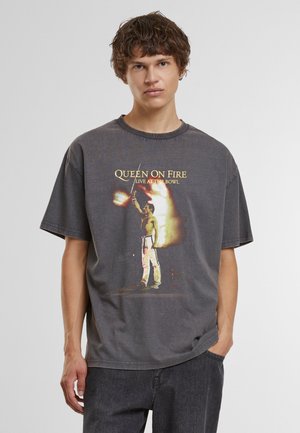 Junger Mann mit braunen lockigen Haaren trägt ein lockeres dunkelgraues T-Shirt mit einer flammenden Figur und Text "Queen On Fire Live At The Bowl."