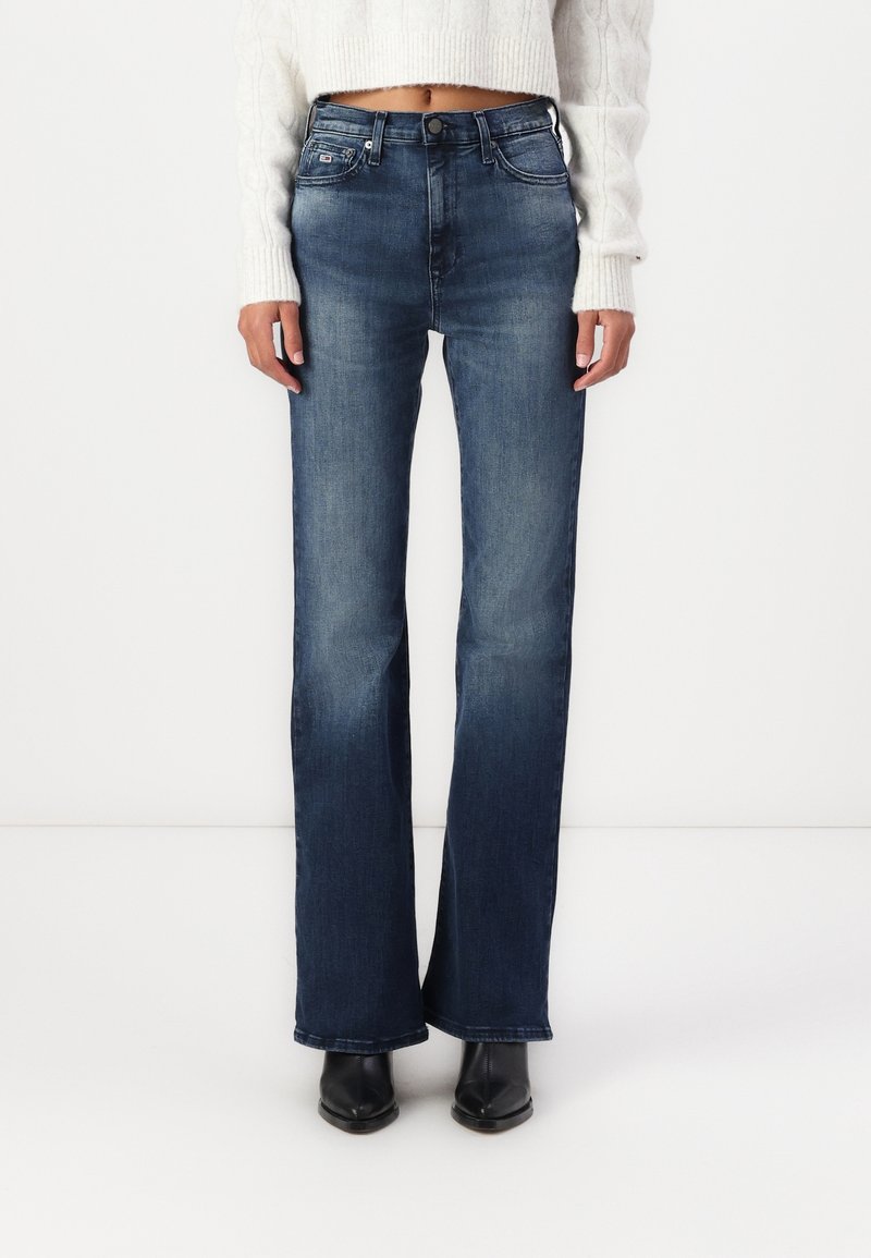Personne portant un jean bootcut bleu foncé taille haute, un pull court blanc et des bottes noires à bout pointu sur fond blanc uni.