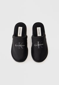 Pár čiernych slip-on papúč Calvin Klein s odpruženou podrážkou a logom vytlačeným na vrchu, vystavený na bielom pozadí.