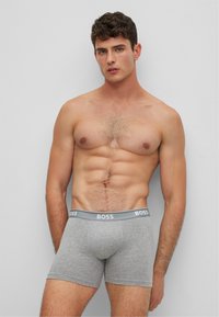 Graue Baumwollboxershorts mit BOSS-Logo am Bund, die einen körpernahen Schnitt und eine glatte Textur aufweisen. Getragen von einem männlichen Model mit muskulösem Körperbau.