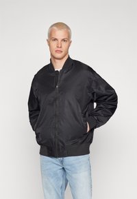 Crna bomber jakna, s patentnim zatvaračem sprijeda, rebrasti završeci i donji rub, glatka satinasta završna obrada, u kombinaciji sa svijetloplavim trapericama. Slim fit, minimalne detalje.
