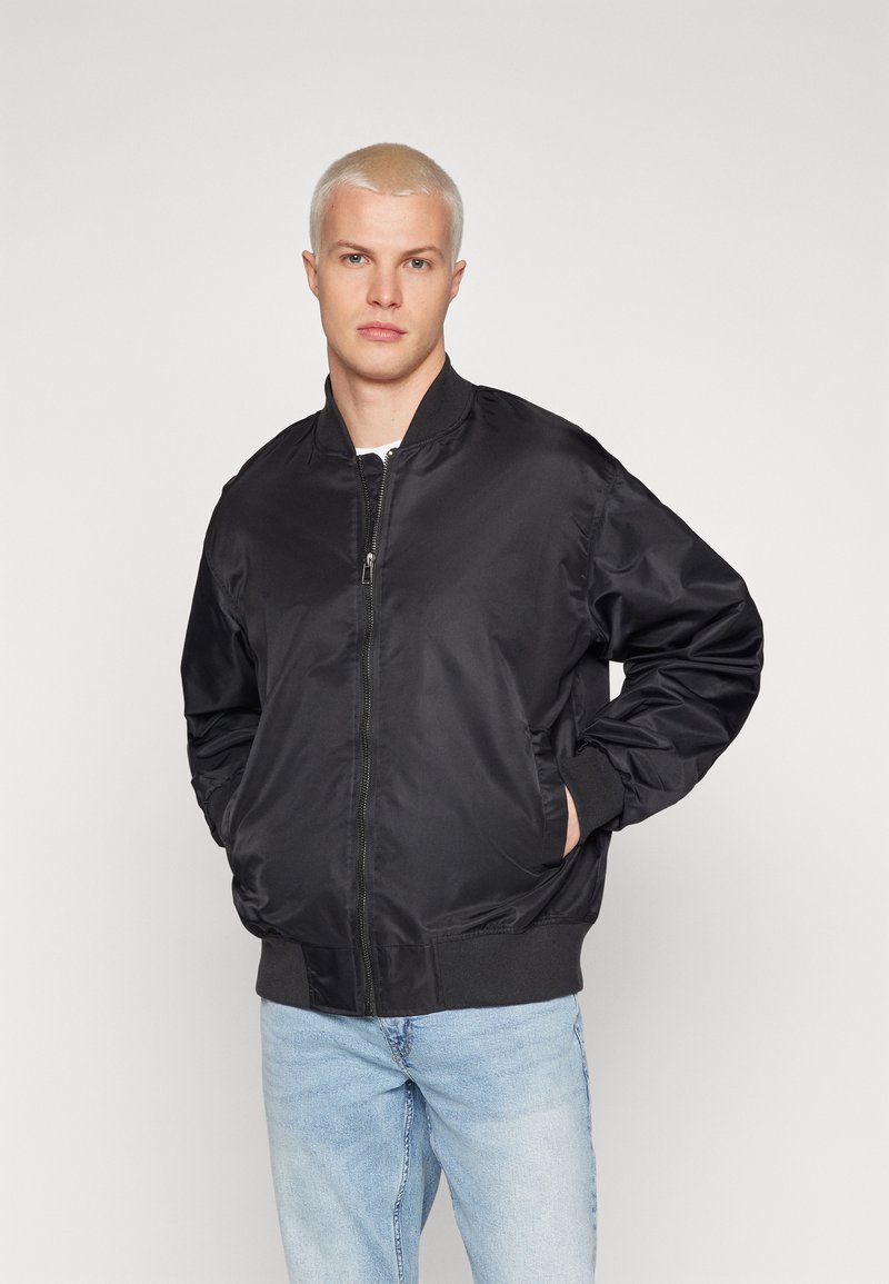 Crna bomber jakna, s patentnim zatvaračem sprijeda, rebrasti završeci i donji rub, glatka satinasta završna obrada, u kombinaciji sa svijetloplavim trapericama. Slim fit, minimalne detalje.