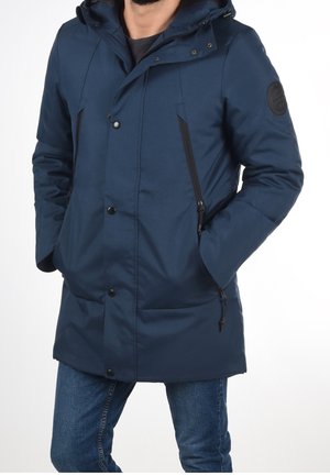 Mann trägt eine marineblaue Kapuzen-Winterjacke mit Reißverschlusstaschen an den Seiten und Druckknöpfen, kombiniert mit blauen Jeans.