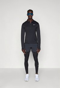 Conjunto desportivo masculino que inclui uma camisola preta de manga comprida, calções cinzentos e leggings pretas. Acessórios incluem óculos de sol azuis e ténis brancos.