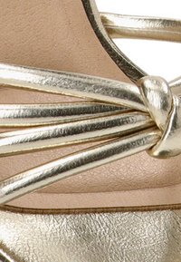 Sandal de tiras metálico dorado con una suave plantilla beige, tiras estrechas con un diseño retorcido y una superficie lisa y reflectante.