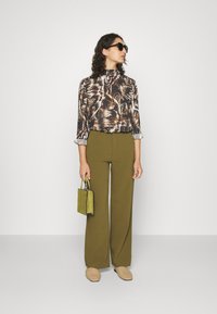 Calças longas de tom verde-oliva com corte ajustado, combinadas com uma blusa escura estampada. Os acessórios incluem uma pequena mala verde brilhante e sapatos de palha em tom bege.