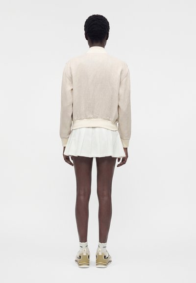 Άτομο με σκούρο δέρμα που φοράει μπεζ μπουφάν, λευκή πλισέ φούστα, λευκές κάλτσες και μπεζ αθλητικά παπούτσια, στέκεται γυρισμένο προς τα πίσω σε μονόχρωμο φόντο.