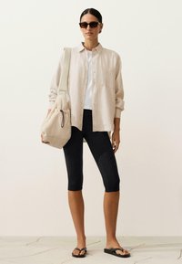 Femeie purtând o cămașă bej supradimensionată cu nasturi, top alb, leggings negri până la genunchi, șlapi negri, ochelari de soare închiși la culoare și având o geantă bej.