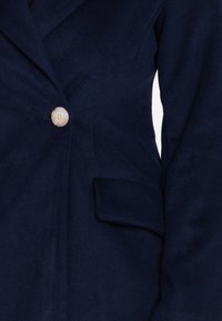 Marineblauer Blazer mit strukturierter Stoffbeschaffenheit, einzelner Holzknopfschlieung und einer Seitentasche, der ein maßgeschneidertes Design hervorhebt.