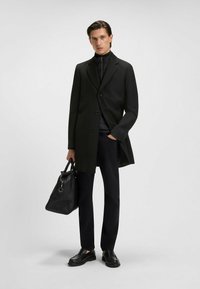 Cappotto nero su misura con colletto a punta, design a un bottone, indossato sopra una giacca nera con zip. Abbinato a pantaloni neri e una borsa di pelle.