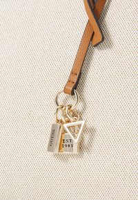 Borsa beige texturizzata con tracolla marrone, caratterizzata da accessori in oro e ciondoli, tra cui un triangolo e un'etichetta incisa "GUESS" e "EST 1981."