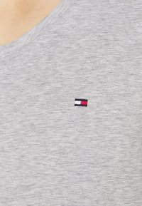 Graues Baumwoll-T-Shirt mit V-Ausschnitt, versehen mit einem kleinen gestickten Logo in Rot, Weiß und Blau auf der linken Brustseite.