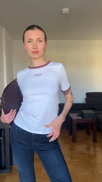 Νεαρή γυναίκα με λευκό μπλουζάκι και μπλε τζιν κρατάει ένα σκούρο πλεκτό φάκελο, ποζάροντας με αυτοπεποίθηση σε ένα σαλόνι.