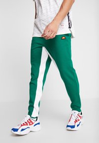 Man som har på sig gröna och vita träningsoverallbyxor med orange logotyp och vita sneakers med blå och röda detaljer, står mot en vit bakgrund.