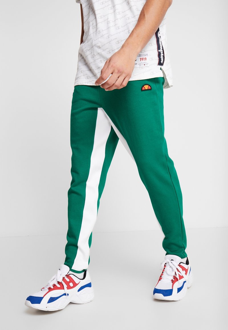 Man som har på sig gröna och vita träningsoverallbyxor med orange logotyp och vita sneakers med blå och röda detaljer, står mot en vit bakgrund.