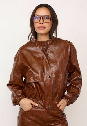 Junge Frau mit langen dunklen Haaren trägt eine braune Lederjacke und Hose, posiert mit den Händen in den Jackentaschen und großen braunen Brillengestellen.